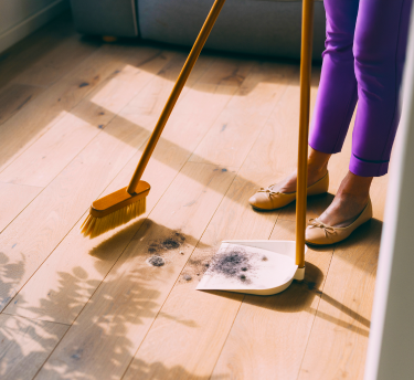 A person sweeps dust and debris from a hardwood floor using a broom and dustpan, suggesting dust an allergy trigger.