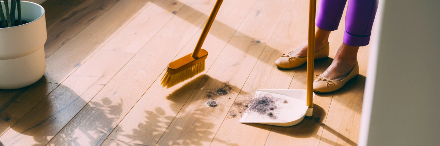 A person sweeps dust and debris from a hardwood floor using a broom and dustpan, suggesting dust an allergy trigger.