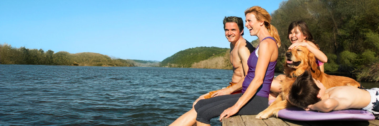 A happy family of two adults and two children sits on a dock by a lake, enjoying an allergy free sunny day.